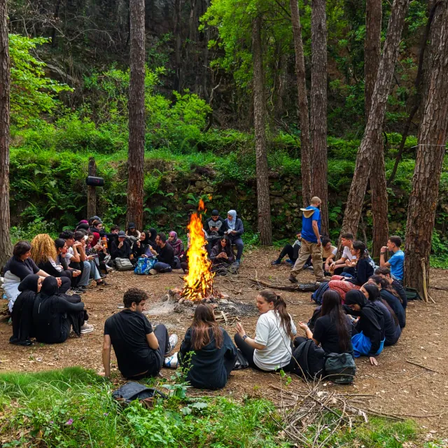 A large group sitting in a circle around a bonfire in a pine clearing.
