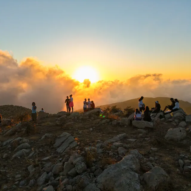 Hikers standing in silhouette on a rocky summit as the sun sets over a sea of cloud.